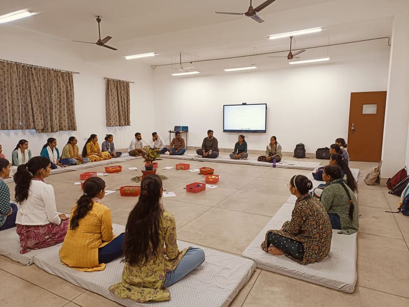 About twenty-five educators sitting in a wide circle on floor mattresses during a residential block at Baghpat.