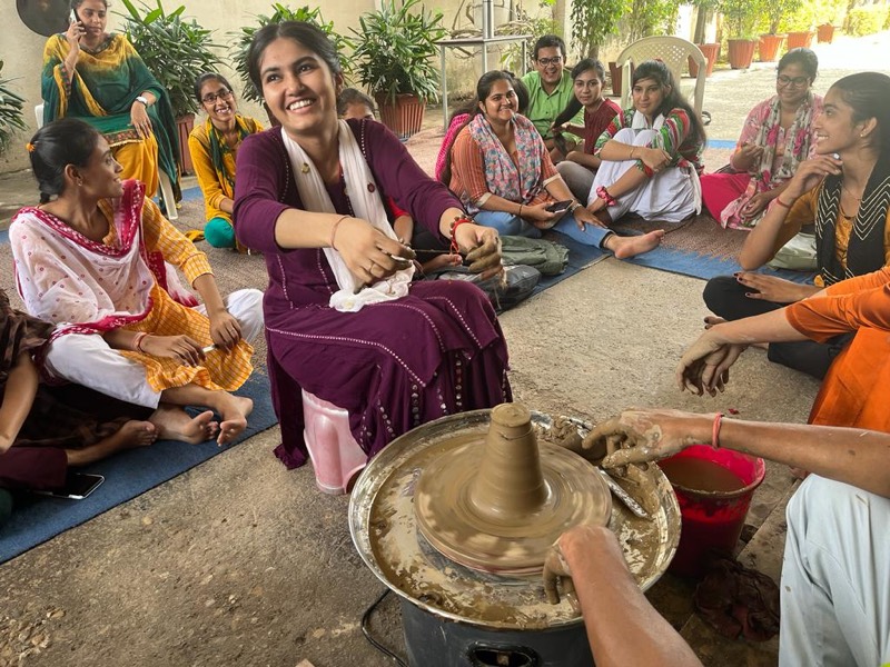A student laughing as she works at a potter’s wheel, watched by a circle of classmates seated on the floor around her.