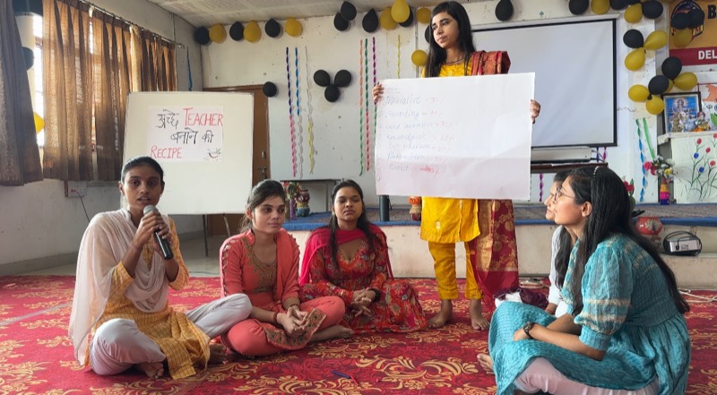 Students seated on the floor presenting a hand-lettered chart headed ‘अच्छे teacher बनने की recipe’ to their classmates.