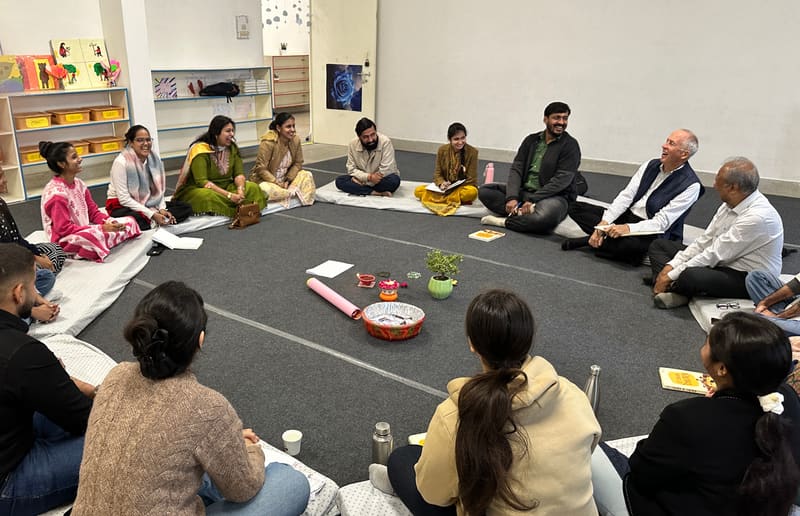 A circle of adults sitting on the floor of a classroom at Kadam, laughing together, with children's artwork and low bookshelves behind them.